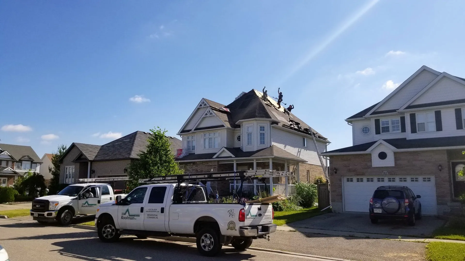 The image shows several people on a house roof under a clear blue sky, working on roofing with parked vehicles from a roofing company below.