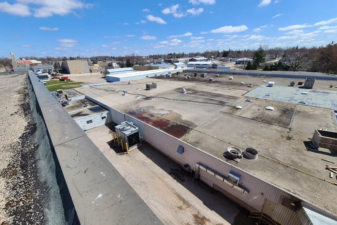 A wide-angle view of industrial rooftops under a clear sky, with HVAC units, vents, stains, and some surrounding buildings and trees visible.