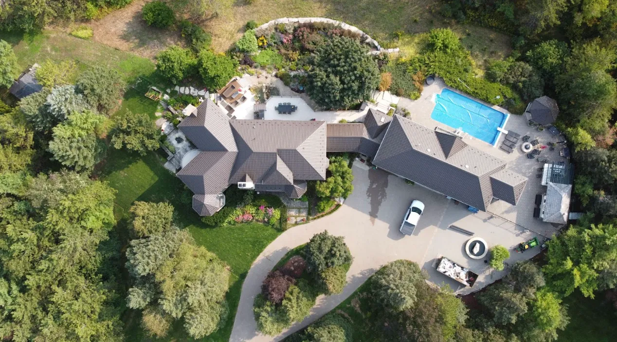 Aerial view of a large residential property with multiple roofs, an outdoor pool, lush gardens, vehicles, and well-manicured lawns surrounded by trees.