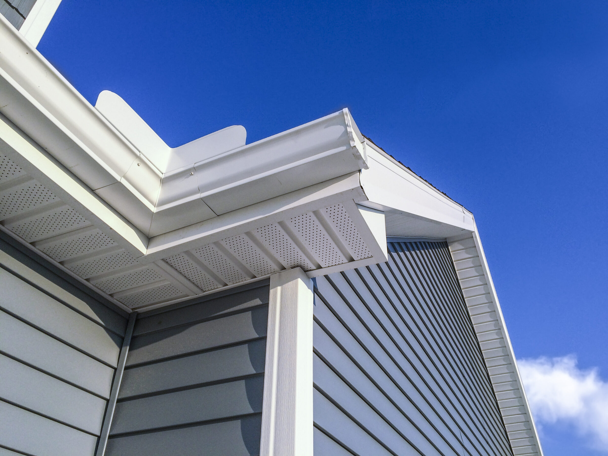 The image shows a close-up of a house's white siding, soffit, and fascia under a clear blue sky, emphasizing architectural details and clean lines.