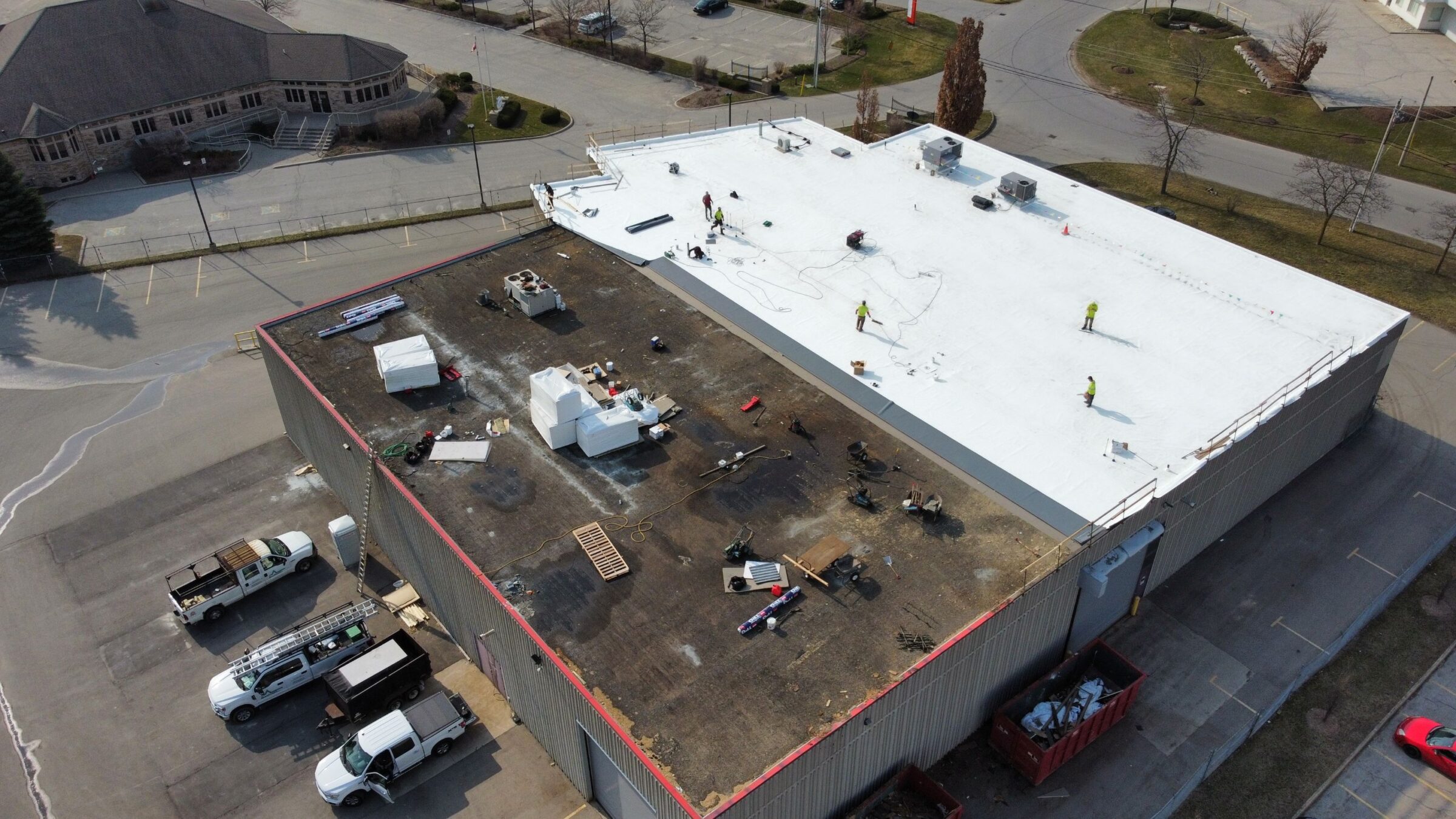 Aerial view of a commercial building with a flat roof where several people are working, vehicles parked below, construction materials scattered, and nearby trees.