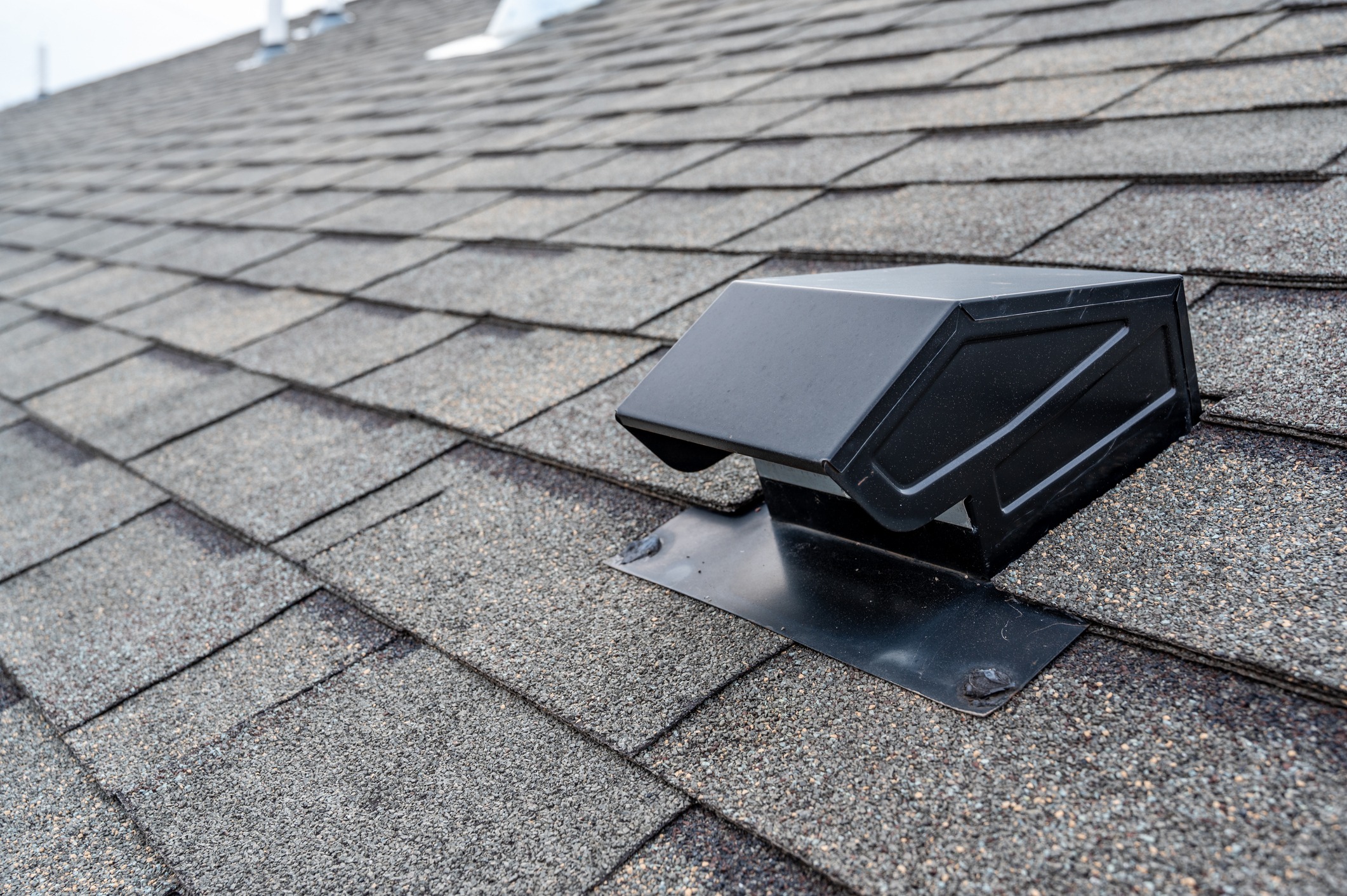 This image shows a close-up of a black roof vent on a shingled rooftop, with a focus on the vent against the textured shingles.