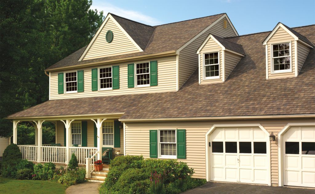 This is a two-story suburban house with beige siding, green shutters, a front porch, white garage doors, and surrounded by trees and a lawn.