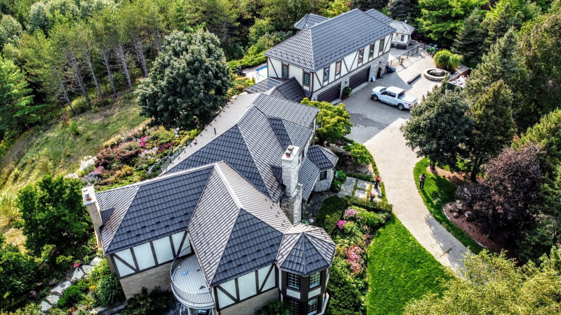 Aerial view of a large, luxurious Tudor-style house with steep gabled roofs, surrounded by lush gardens, trees, a manicured lawn, and a cobblestone driveway.