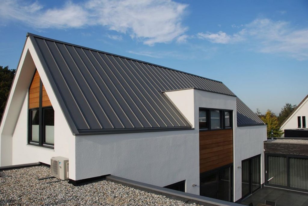 Modern house with a pitched metal roof, white walls, wood accents, large windows, and an outdoor unit, set against a clear blue sky.