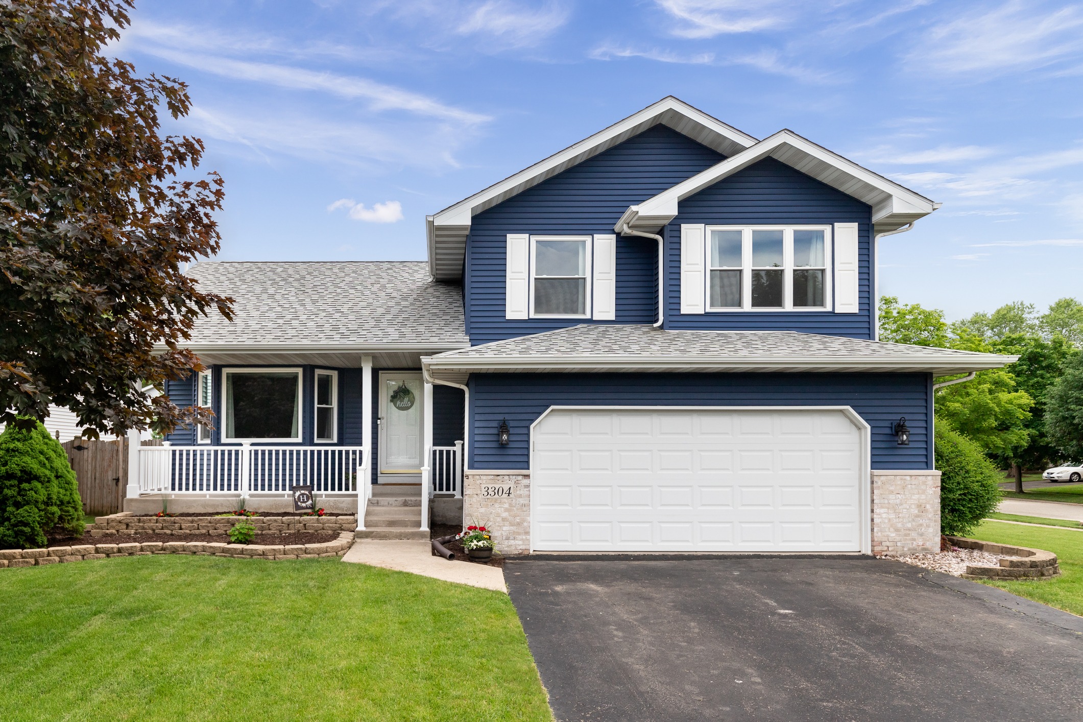 A two-story suburban house with blue siding, white trim, a double garage door, and a well-maintained lawn under a clear sky.
