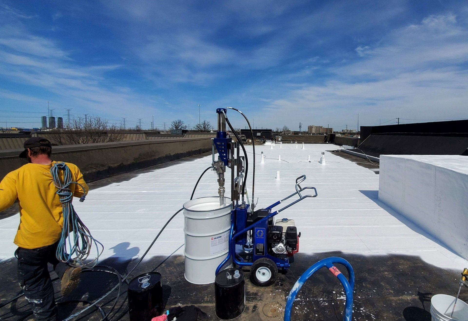 A person works on a flat rooftop coated with white material, near industrial equipment and paint containers, under a clear blue sky. Urban skyline in distance.