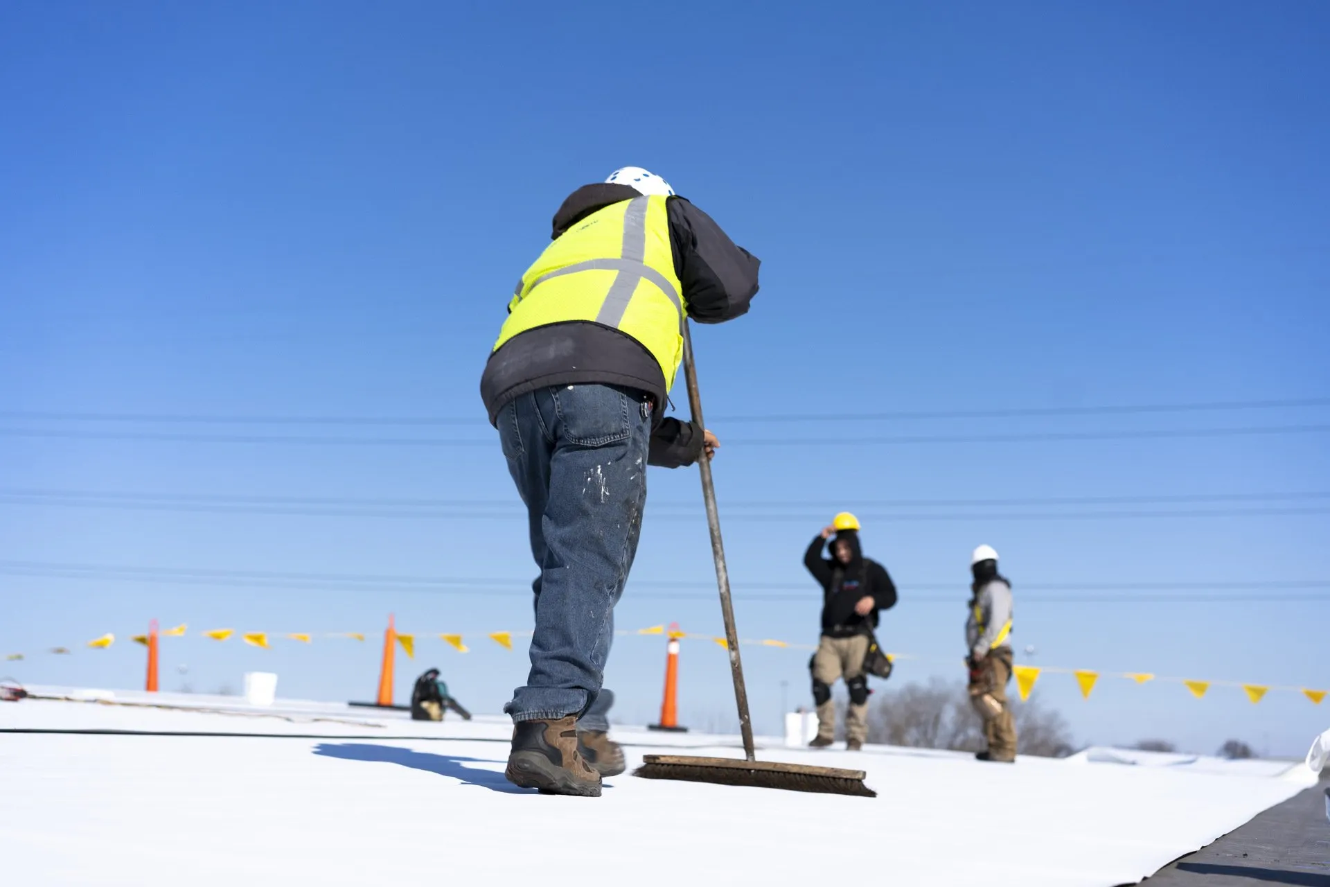 A person in a high-visibility vest and hard hat works on a construction site with a tool, alongside colleagues, under a clear blue sky.