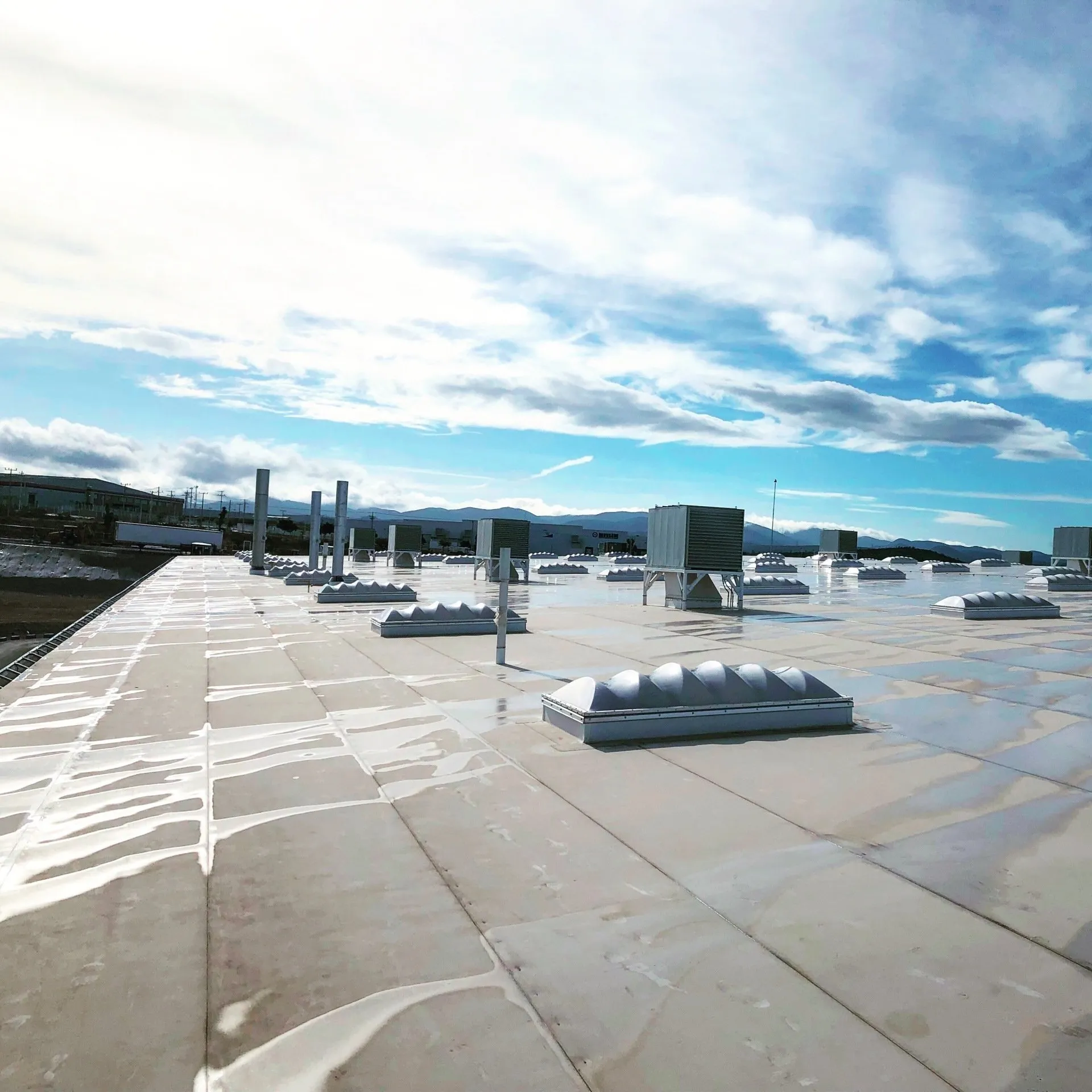 The image shows a flat rooftop with rows of air vents under a clear blue sky with scattered clouds. Wet surfaces suggest recent rain.