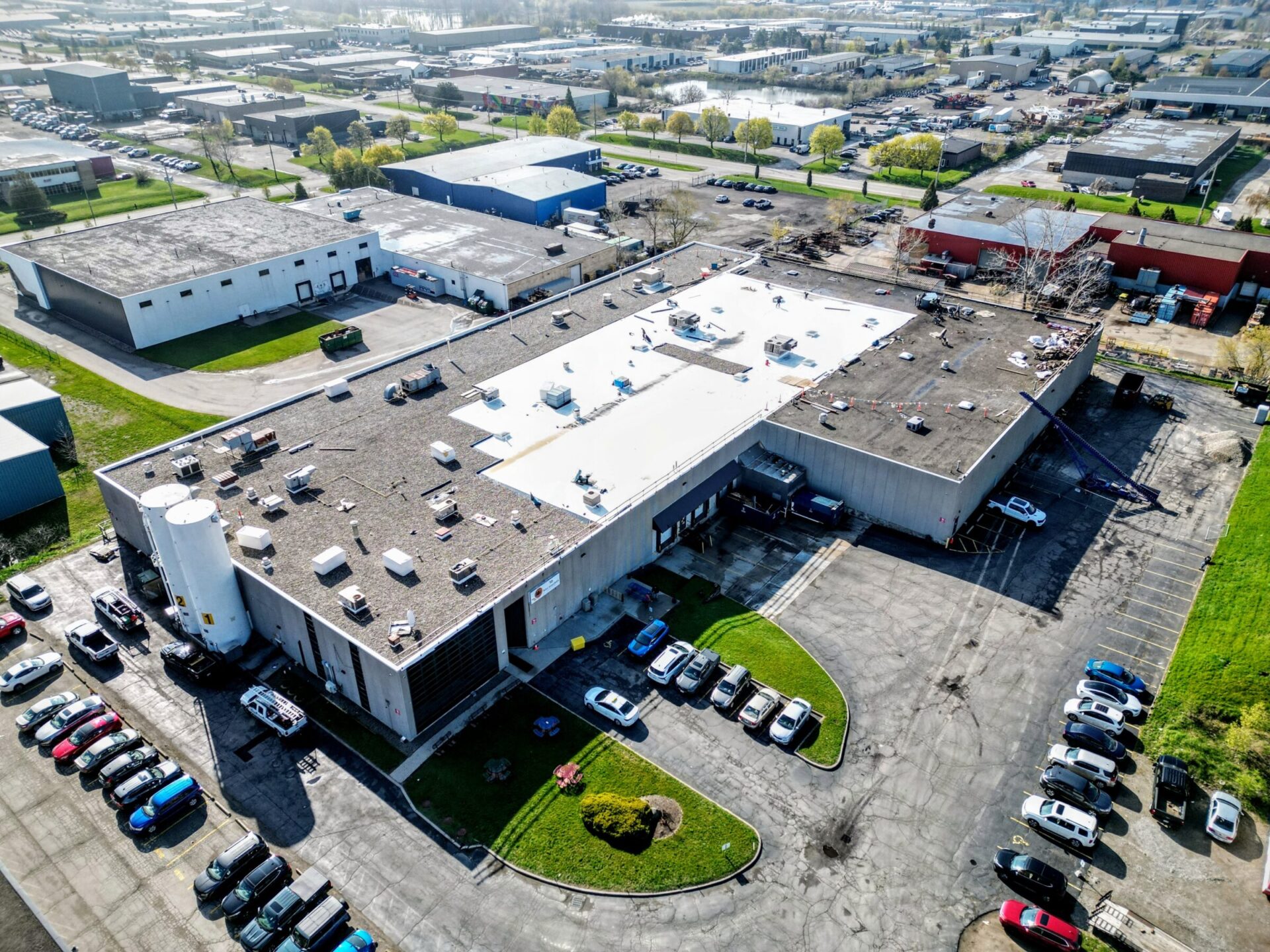 Aerial view of an industrial area with warehouses, parked cars, and a crane on a rooftop under a clear blue sky. Some trees and grass visible.