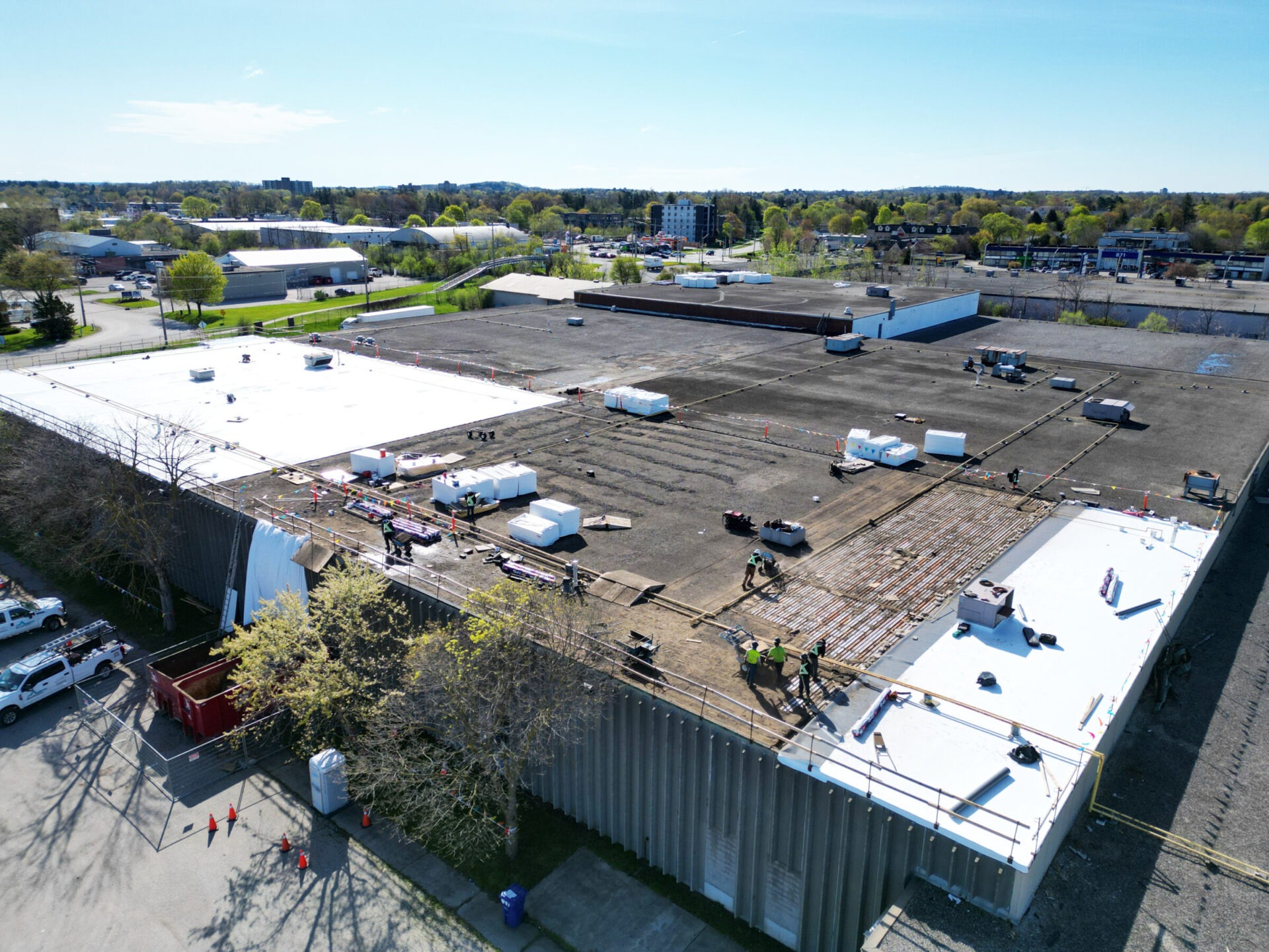 An aerial view of a spacious industrial site with large buildings, parking areas, several vehicles, and people working on a rooftop construction project.