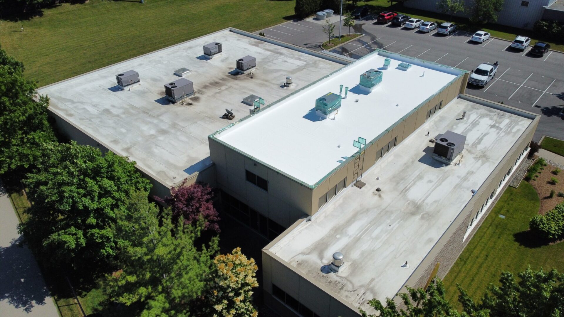 An aerial view of a flat-roofed commercial building with HVAC units, surrounded by a parking lot, trees, and well-maintained grassy areas on a sunny day.