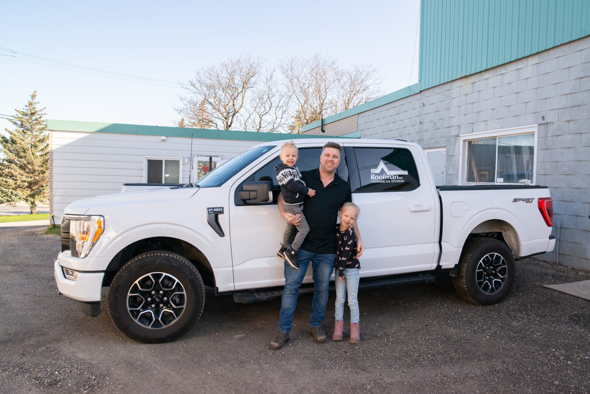 A person is holding a child next to a pickup truck with another child standing beside them, all posing for a photo. They appear happy and outdoors.
