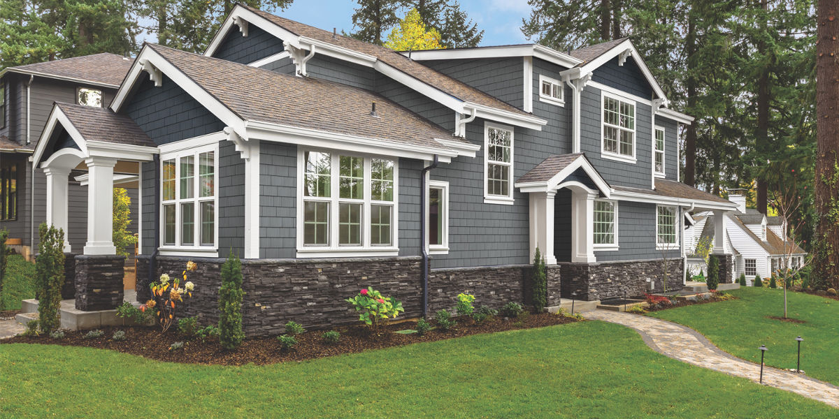 The image features a large two-story house with dark gray siding, white trim, a stone foundation, and a landscaped yard with a walkway.