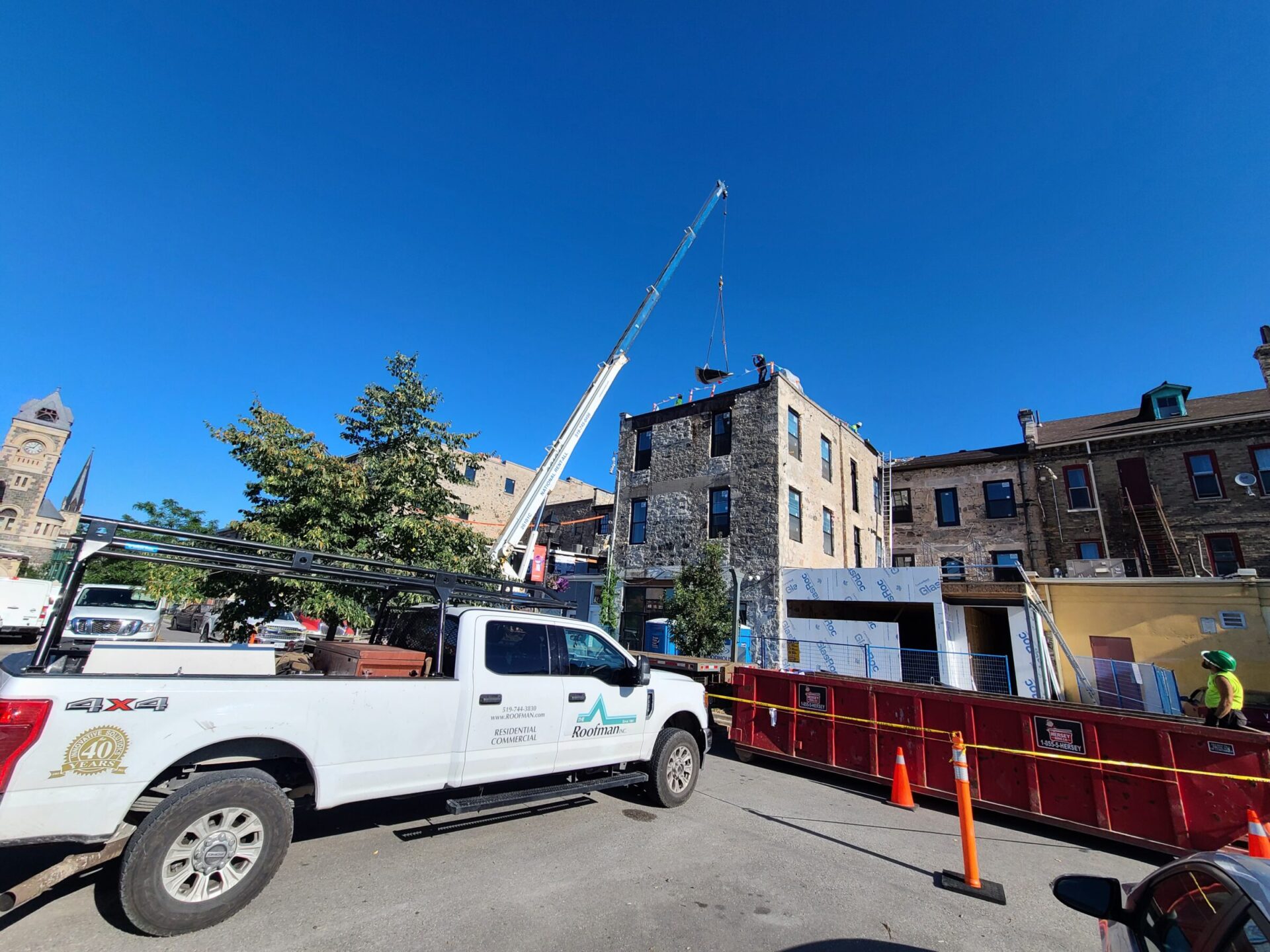 A construction scene with a crane lifting materials, a truck parked nearby, and a person in a yellow vest observing, set against a clear blue sky.