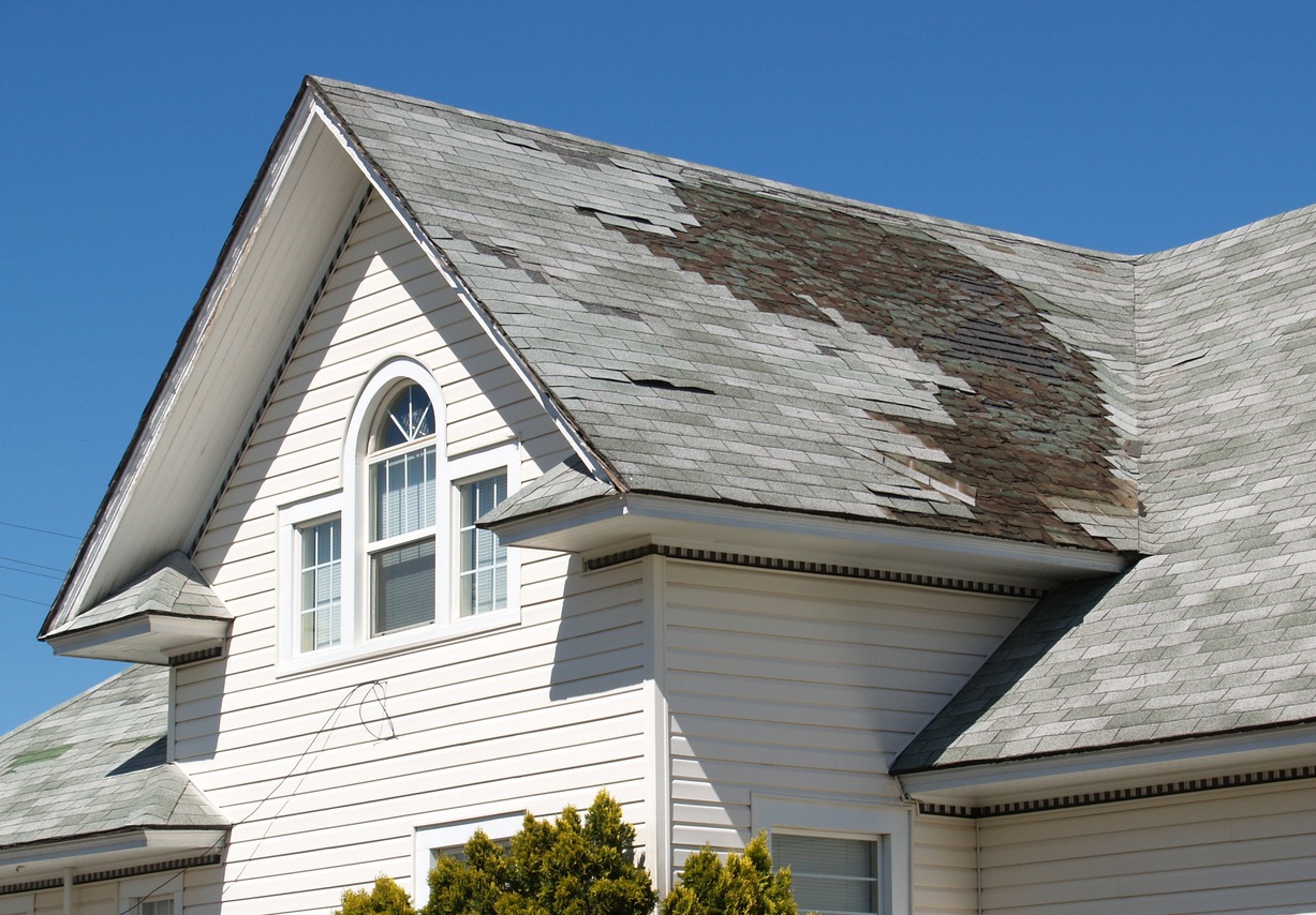 A weathered house roof with several shingles missing and damaged, suggesting neglect or disrepair, above white siding and a decorative window.