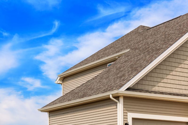 A suburban house with beige siding and a shingled roof under a clear blue sky with wispy clouds, no people visible.