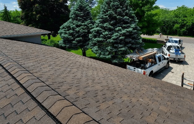 Brown shingled roof with utility trucks parked in driveway. Green trees line the property, and a person stands by a truck under a clear sky.