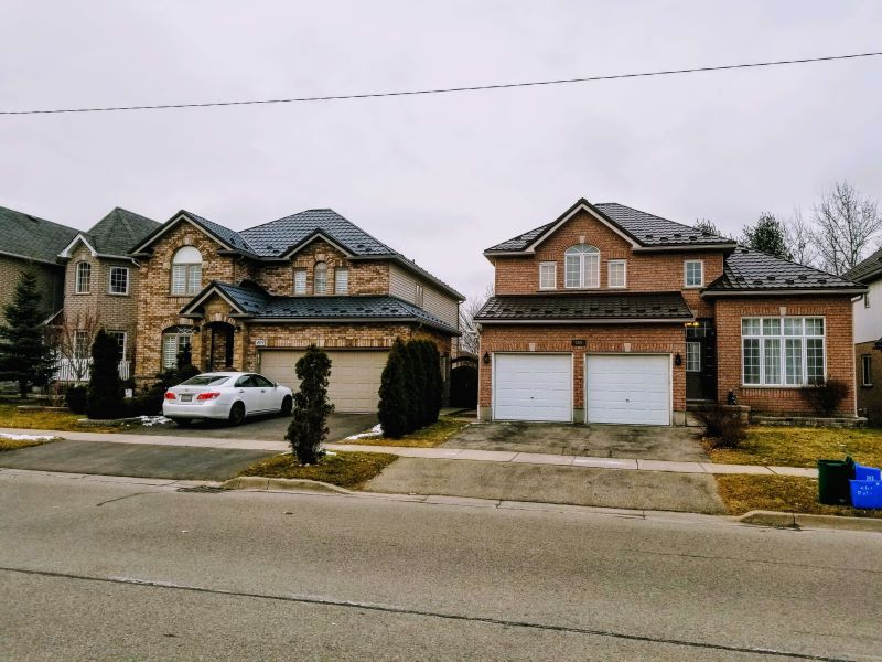 Suburban street with two brick houses, driveways, and a parked car. Overcast sky, leafless trees, green recycling bin on the sidewalk.