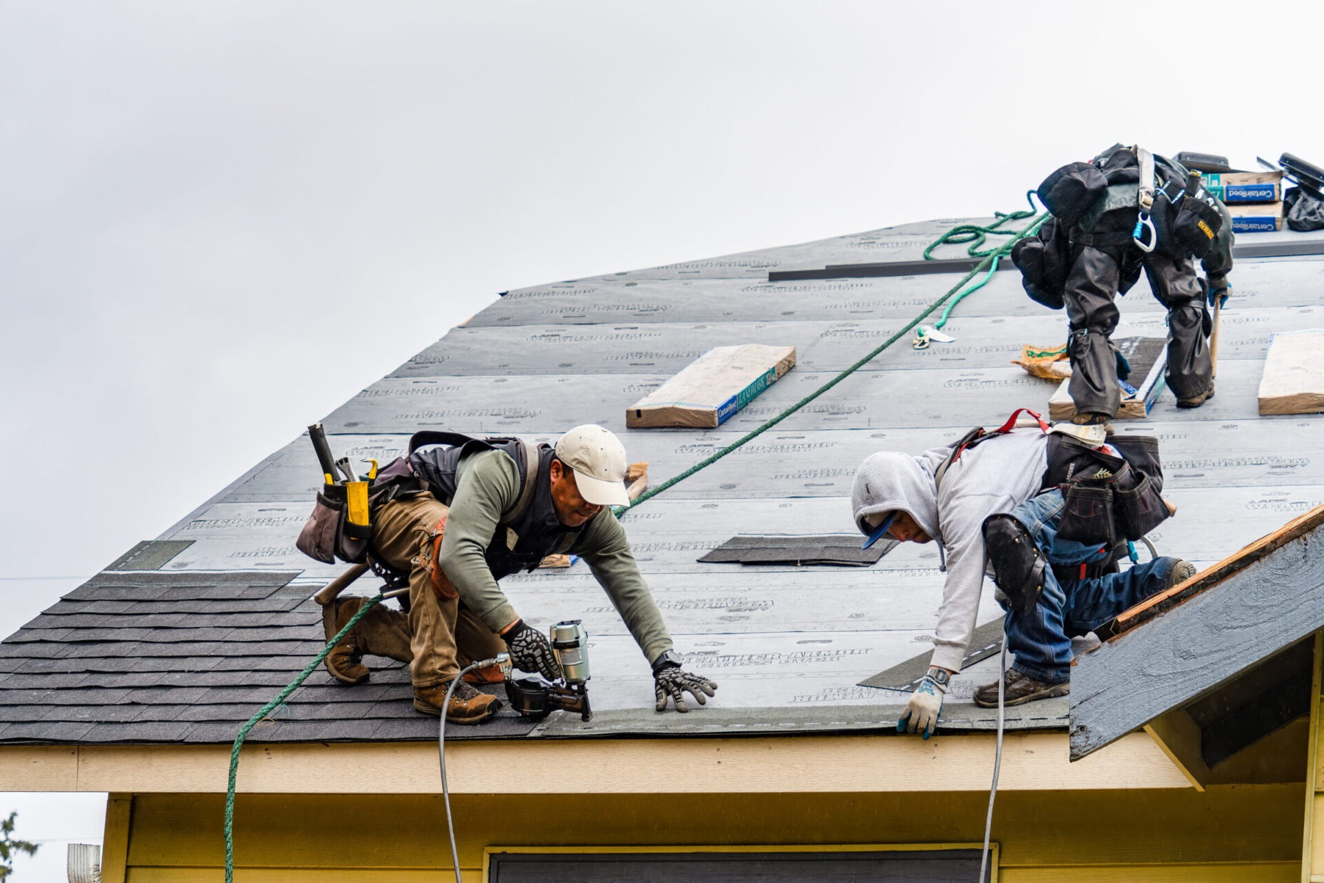 Three people work on a residential roof, installing shingles with various tools and wearing safety gear under overcast skies.