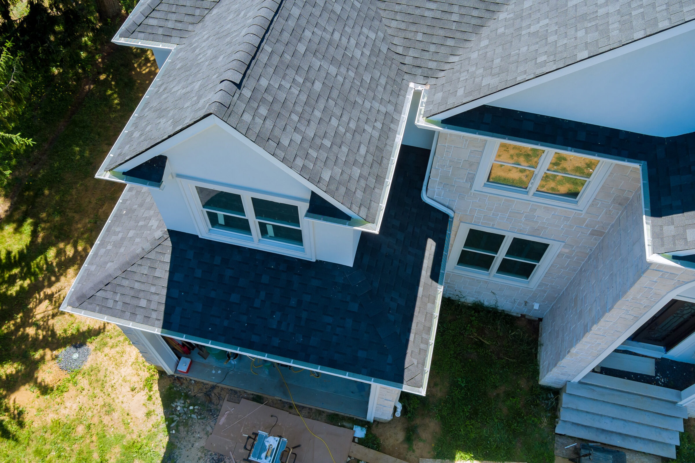 Aerial view of a modern house roof with blue shingles, white walls, and large windows, surrounded by greenery and construction materials below.