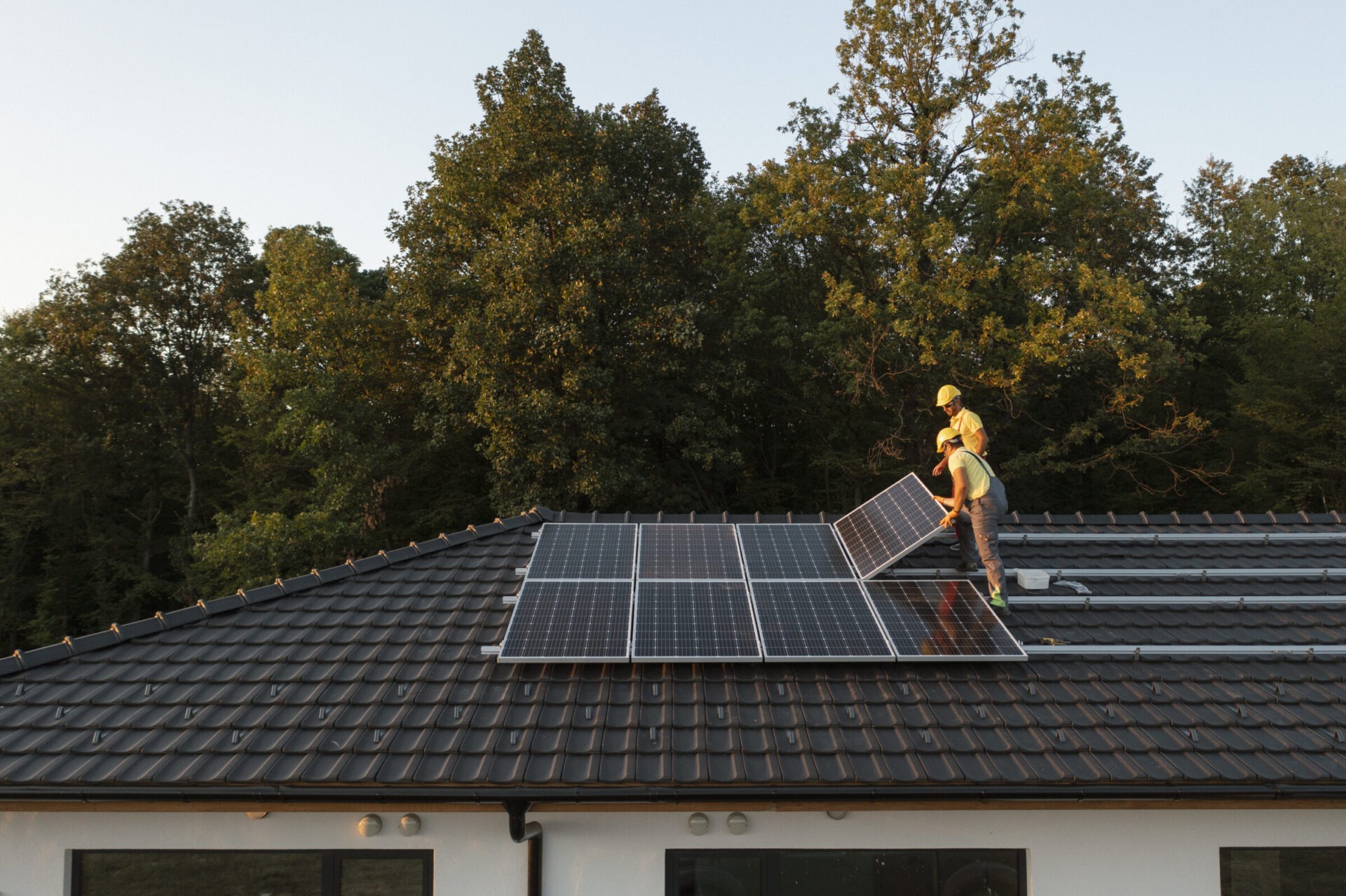 Two people install solar panels on a sloped rooftop surrounded by trees, under a clear sky.