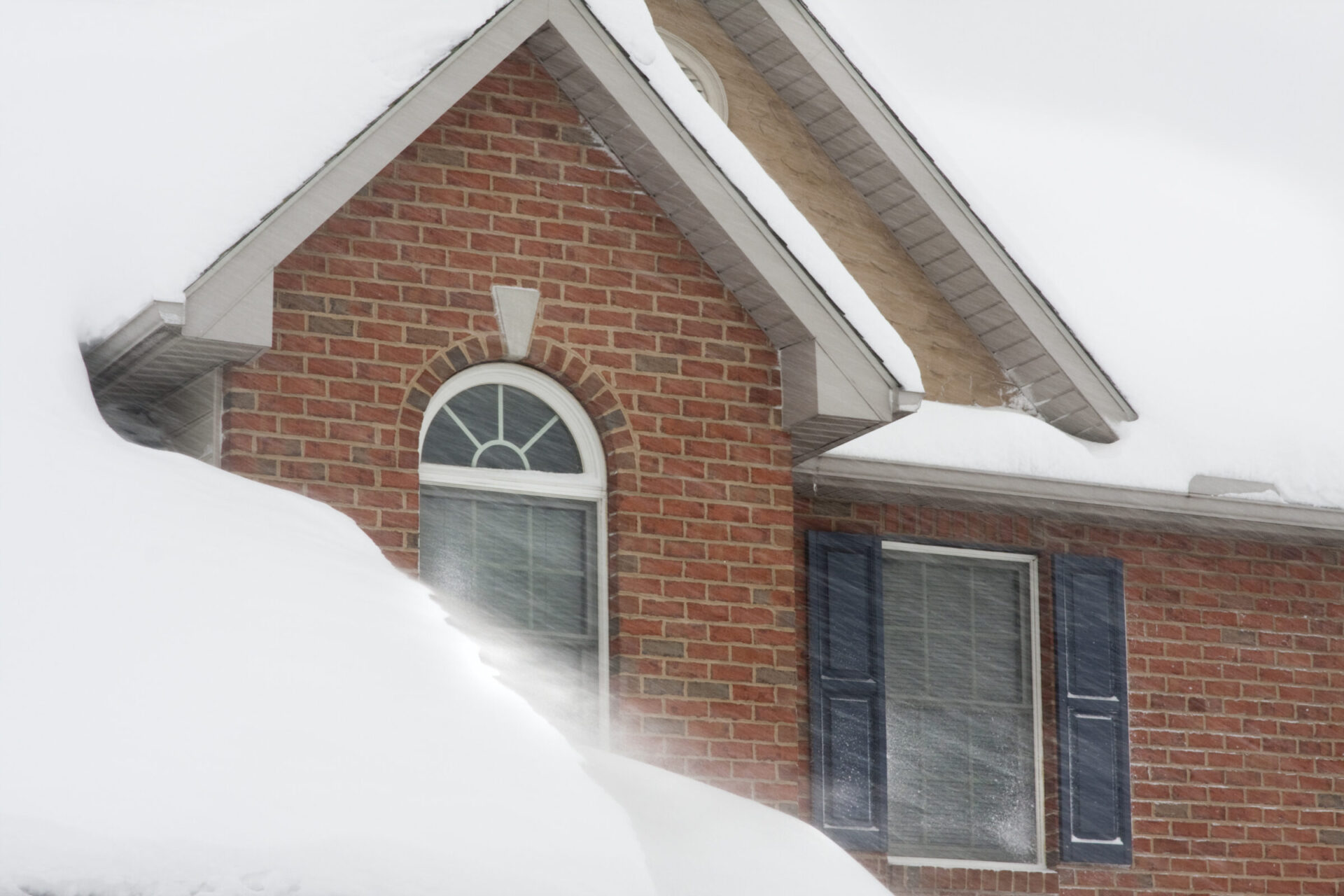 Snow-covered brick house with arched window and shutters, partially obscured by drifting snow. Winter scene with heavy snowfall on the roof.