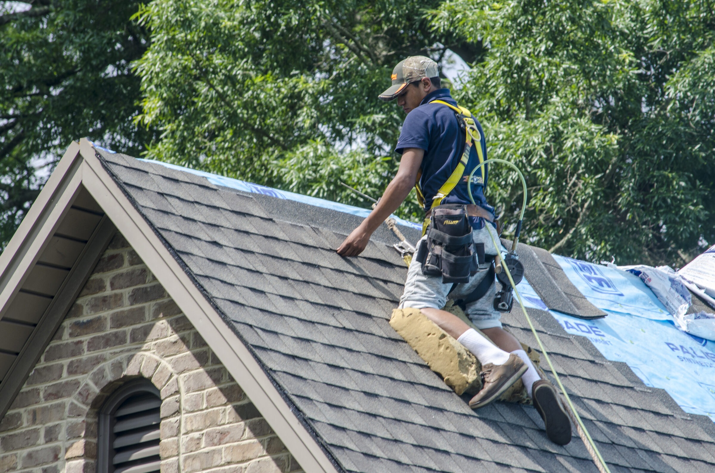 A person in safety harness repairs shingles on a steep roof, surrounded by trees. The house has a brick exterior.