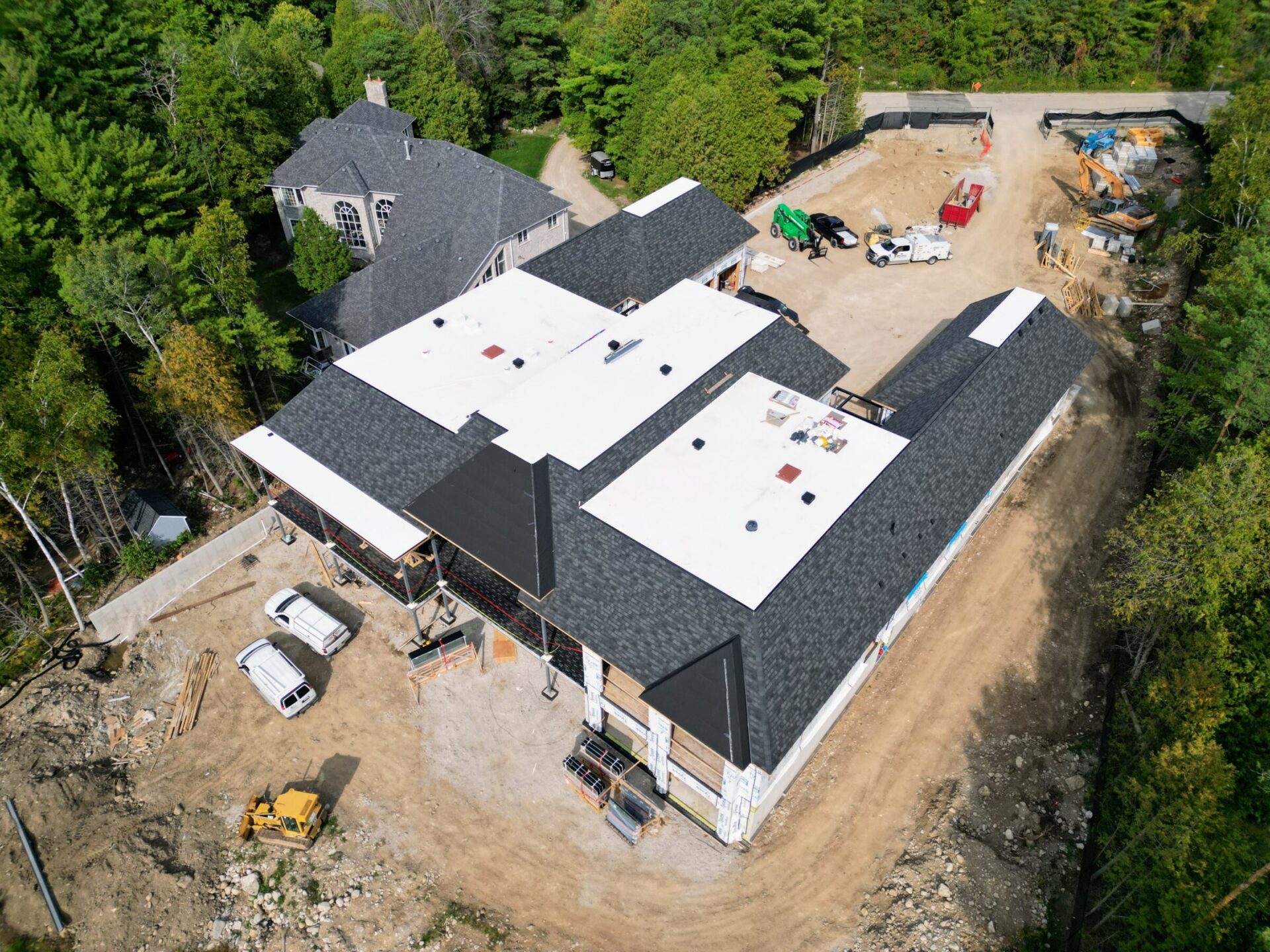 Aerial view of a construction site in a wooded area, featuring a large building, several vehicles, and scattered construction materials.