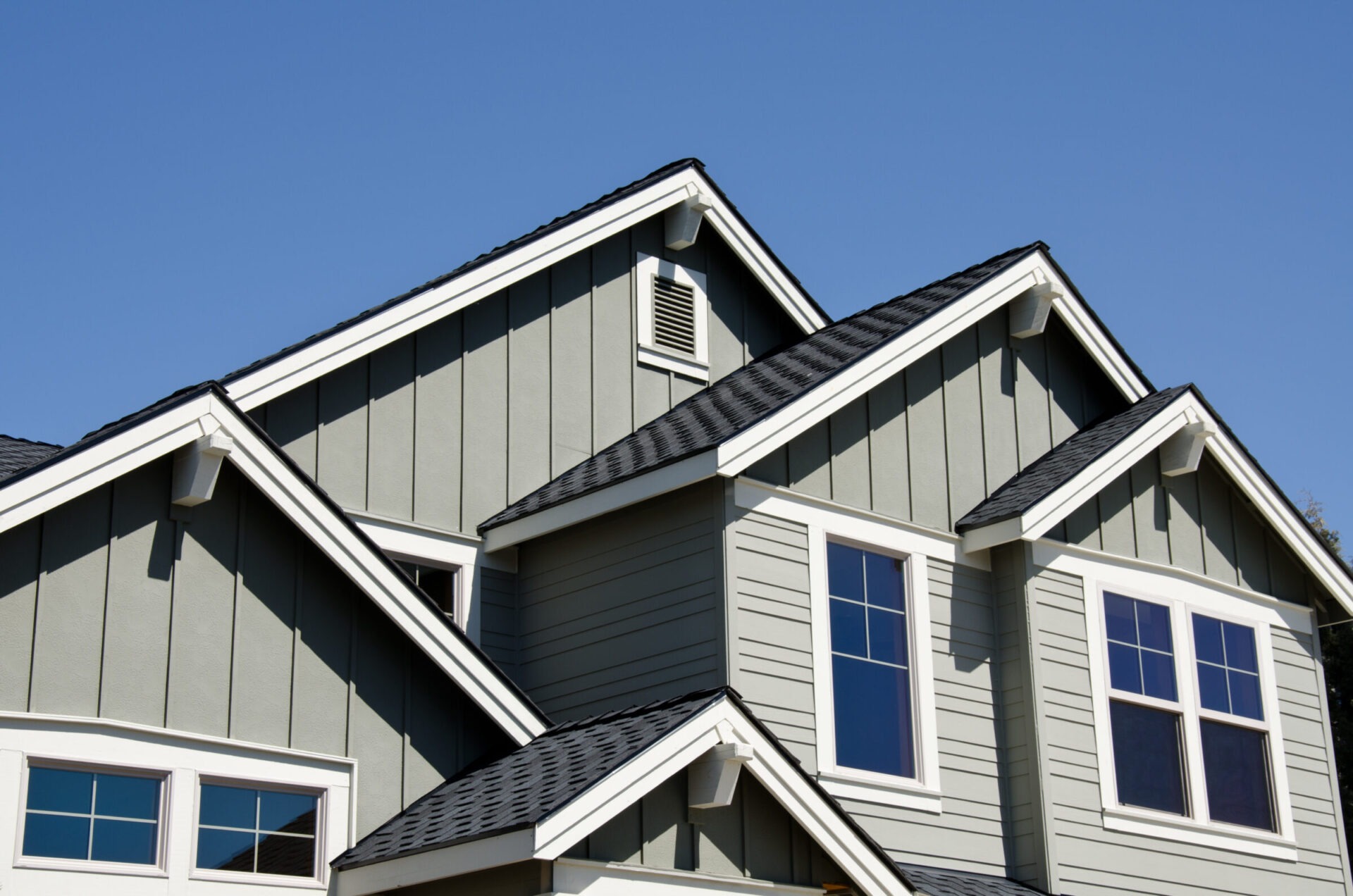 Close-up view of a modern house featuring multiple gabled rooftops with dark shingles, gray board and batten siding, and white trim, all set against a clear blue sky.