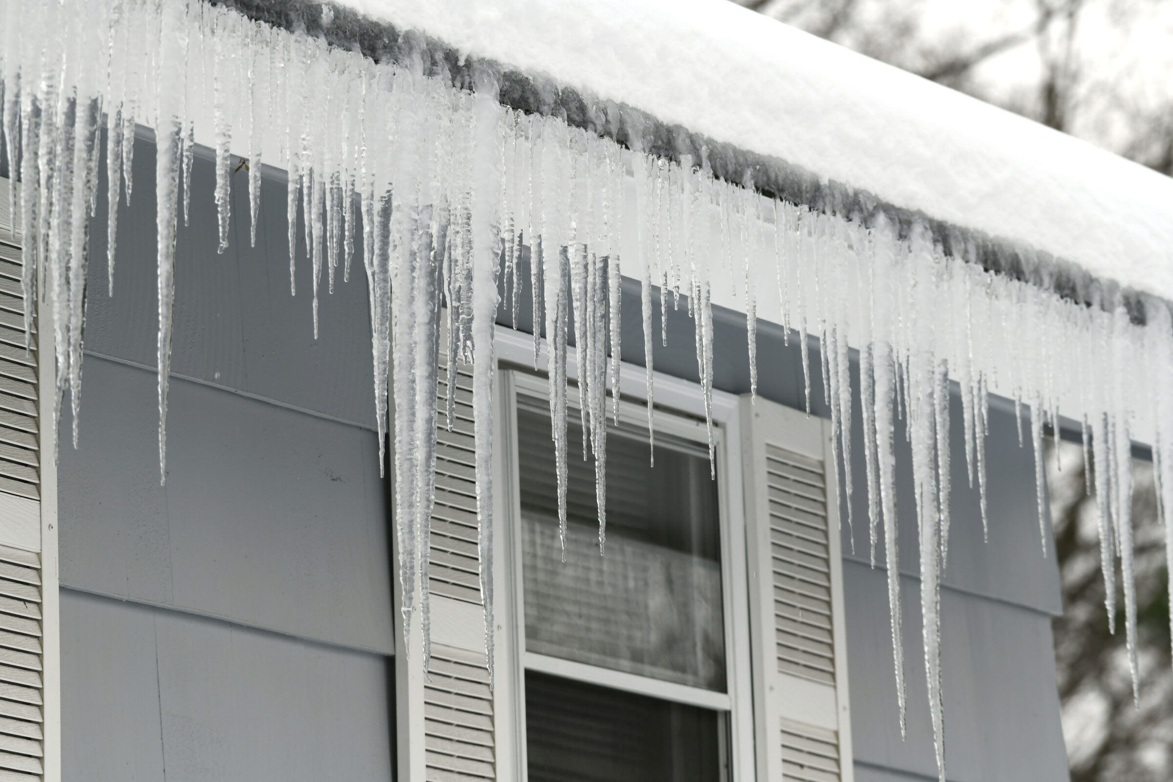 Close-up of a snow-covered roof with long, sharp icicles hanging over the edge above a window framed with white shutters. The icicles vary in length and density, signaling the effects of freezing temperatures and poor roof insulation or gutter drainage.