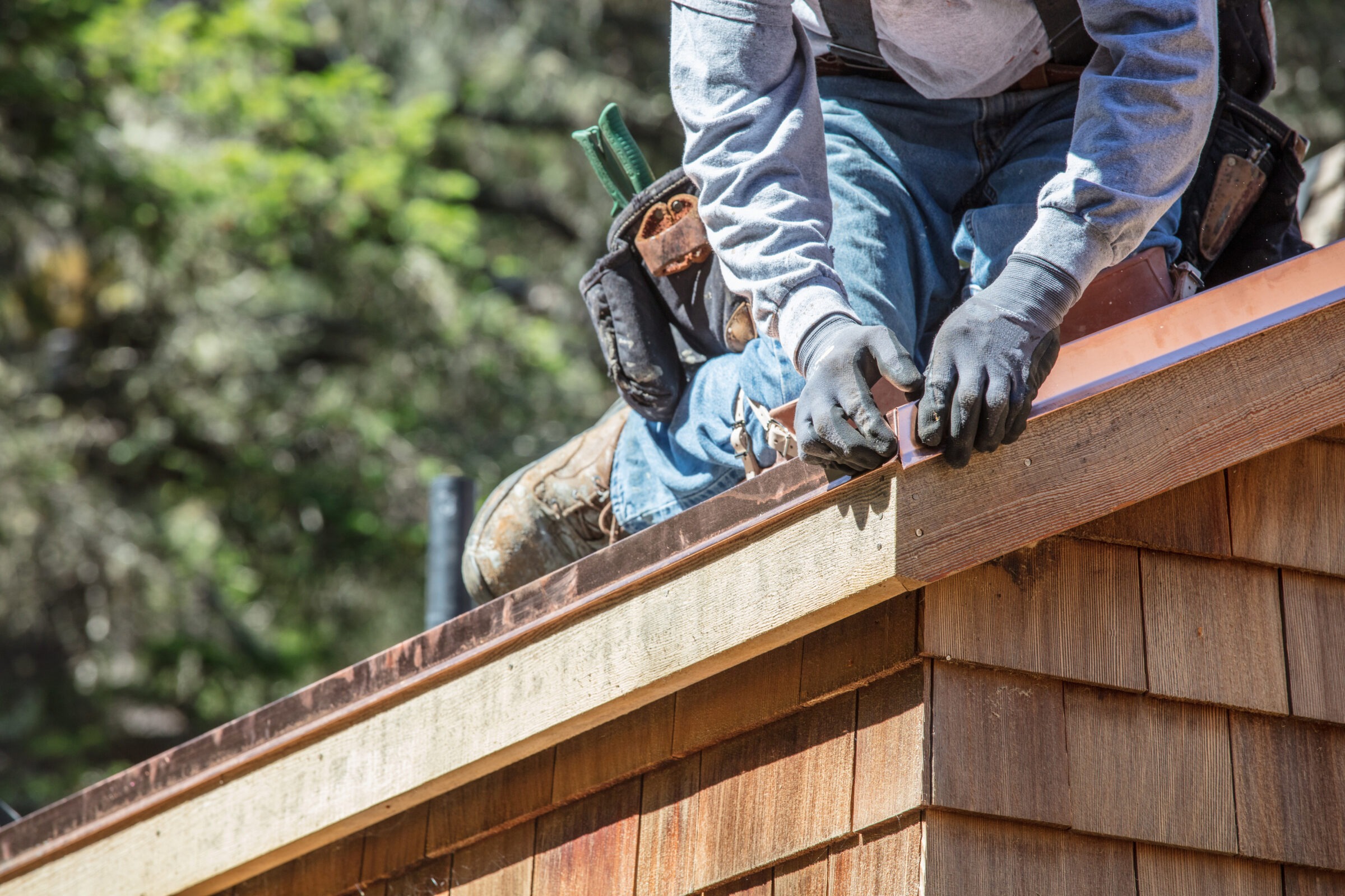 A construction worker installs copper flashing along the edge of a wood-shingled roof, using gloves and a tool belt while kneeling on the structure under sunny daylight.