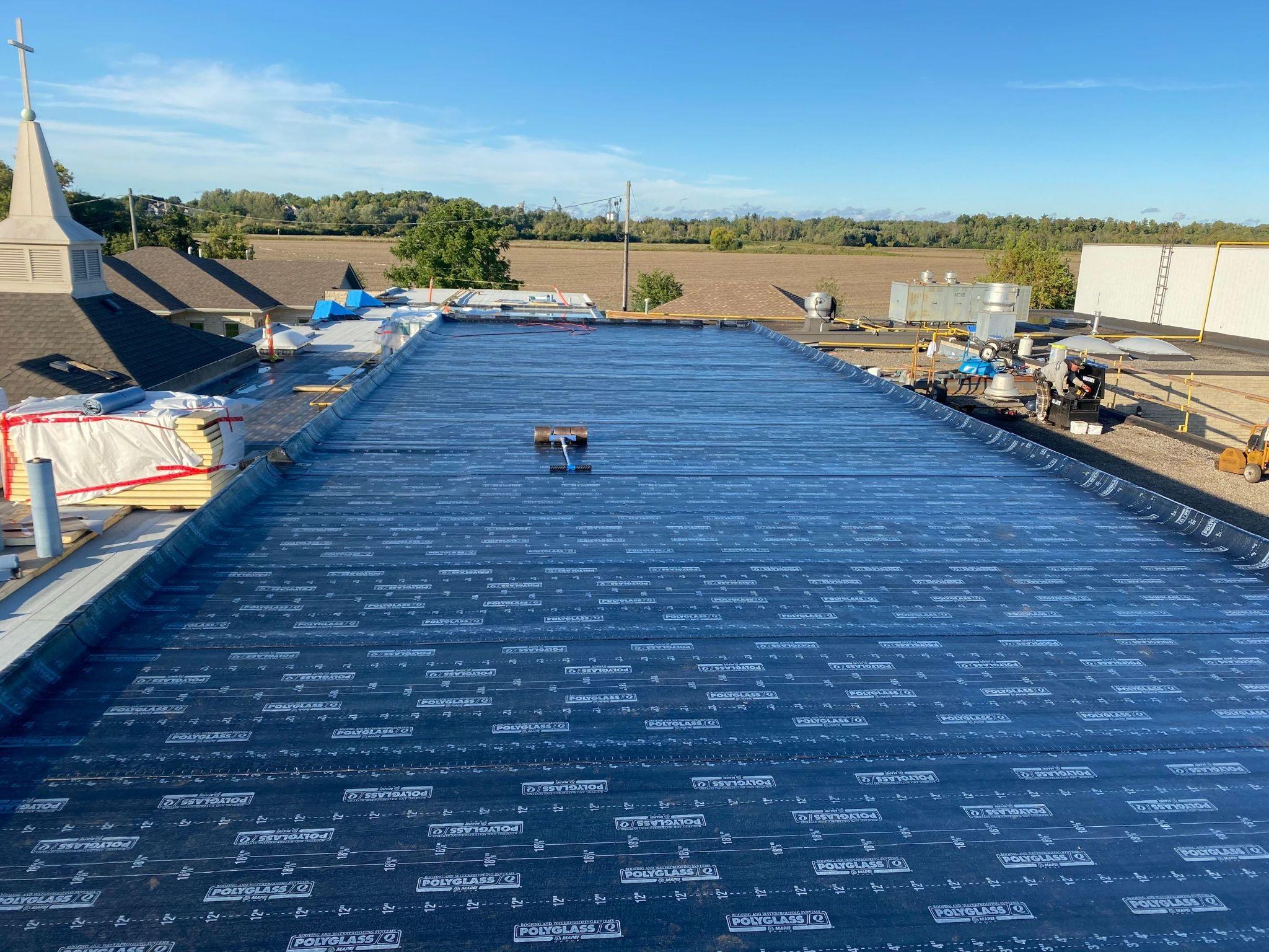 Commercial flat roof under construction with black Polyglass base sheet membrane being installed. Roofing materials and tools are visible across the roof with trees and a church steeple in the background.