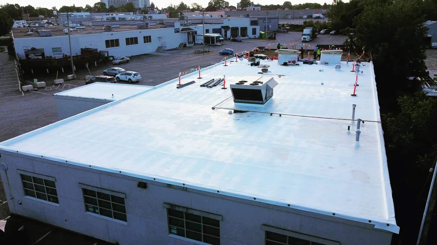 Aerial view of a commercial flat roof installation in progress, covered with a smooth white thermoplastic membrane (TPO). Equipment, materials, and orange safety cones are visible around the rooftop work area.