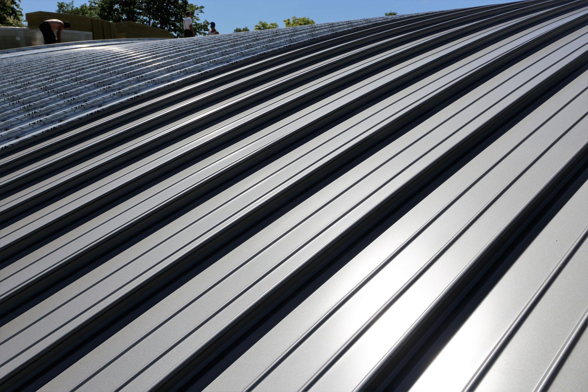 A curved silver standing seam metal roof reflecting sunlight, with workers in the background installing roofing panels.