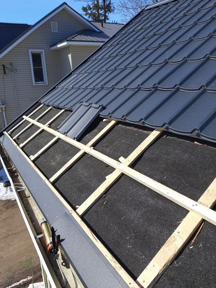 A partially installed dark gray metal tile roof showing the wooden batten framework and black underlayment during construction.