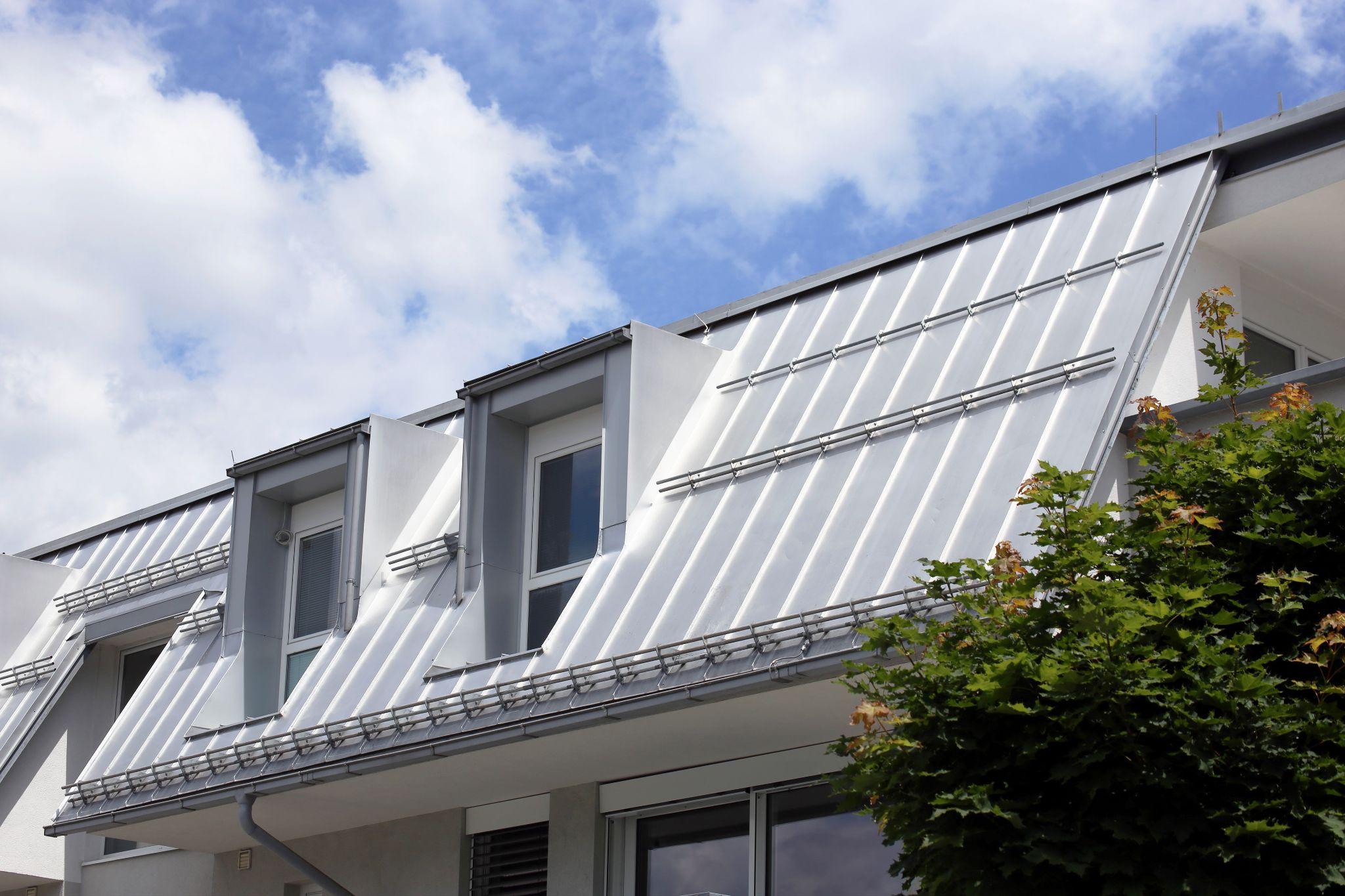 A slanted silver metal roof with dormer windows and integrated gutters on a modern white building beneath a partly cloudy blue sky.
