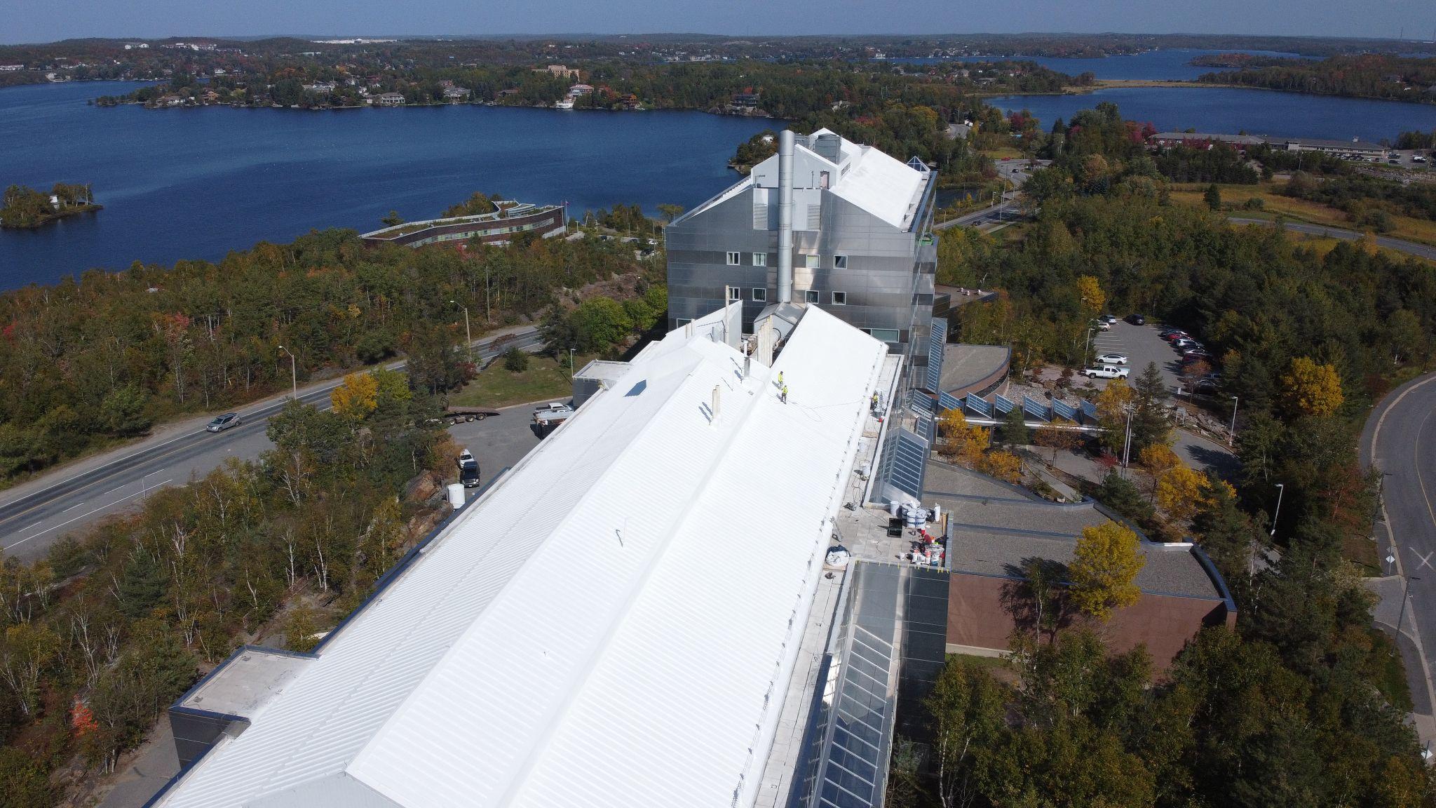 Aerial photo of a large industrial building with a white metal roof beside a road, surrounded by trees and overlooking a lake.