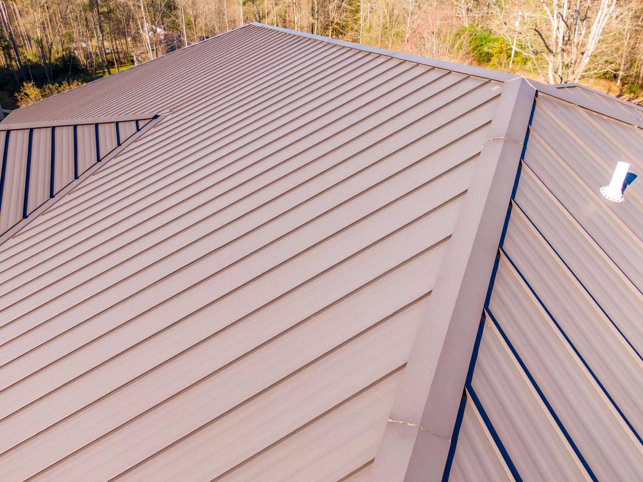 Close-up aerial shot of a brown standing seam metal roof with clean lines and seams, surrounded by bare trees in the background.