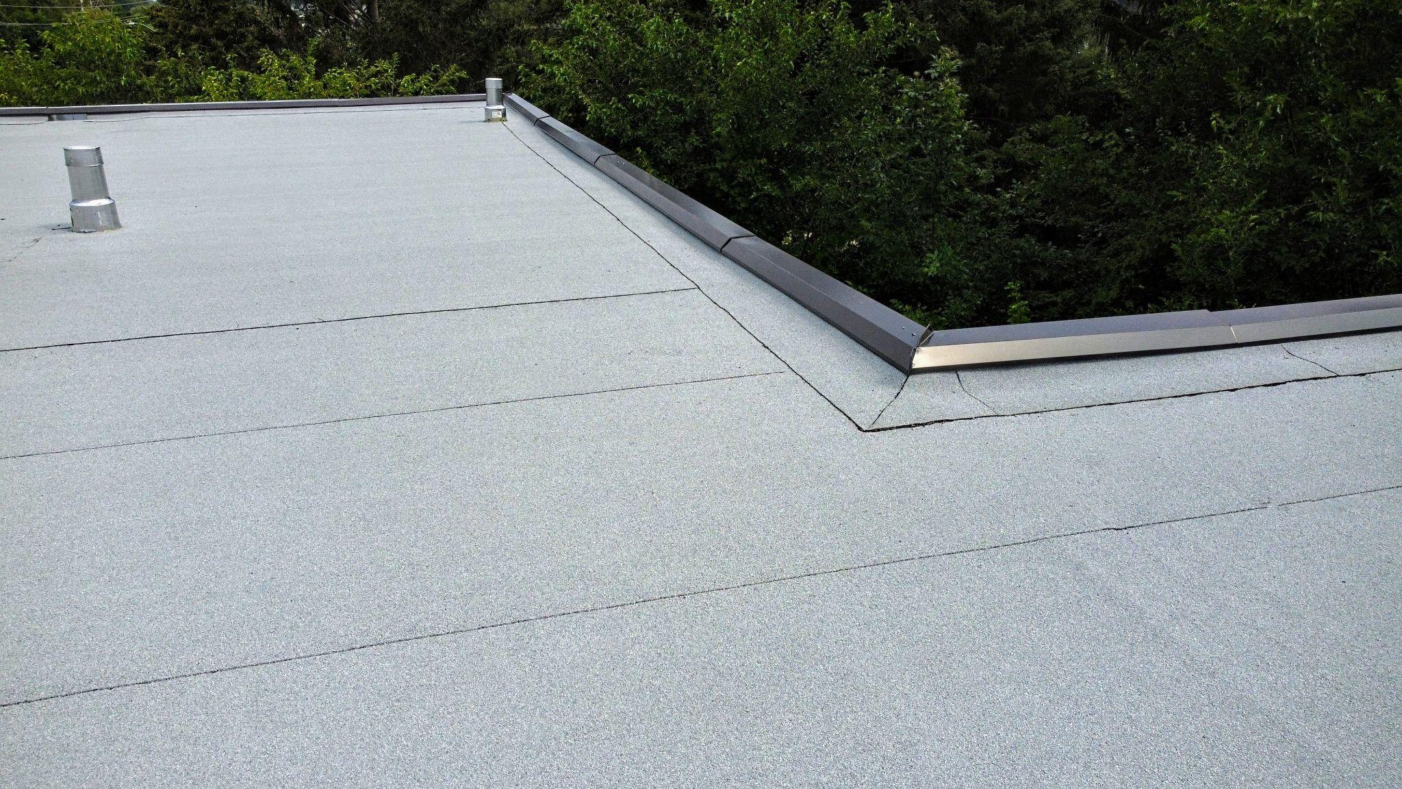 Close-up of a finished flat roof surface with grey modified bitumen membrane, metal flashing, and visible seams near the roof edge bordered by green trees.