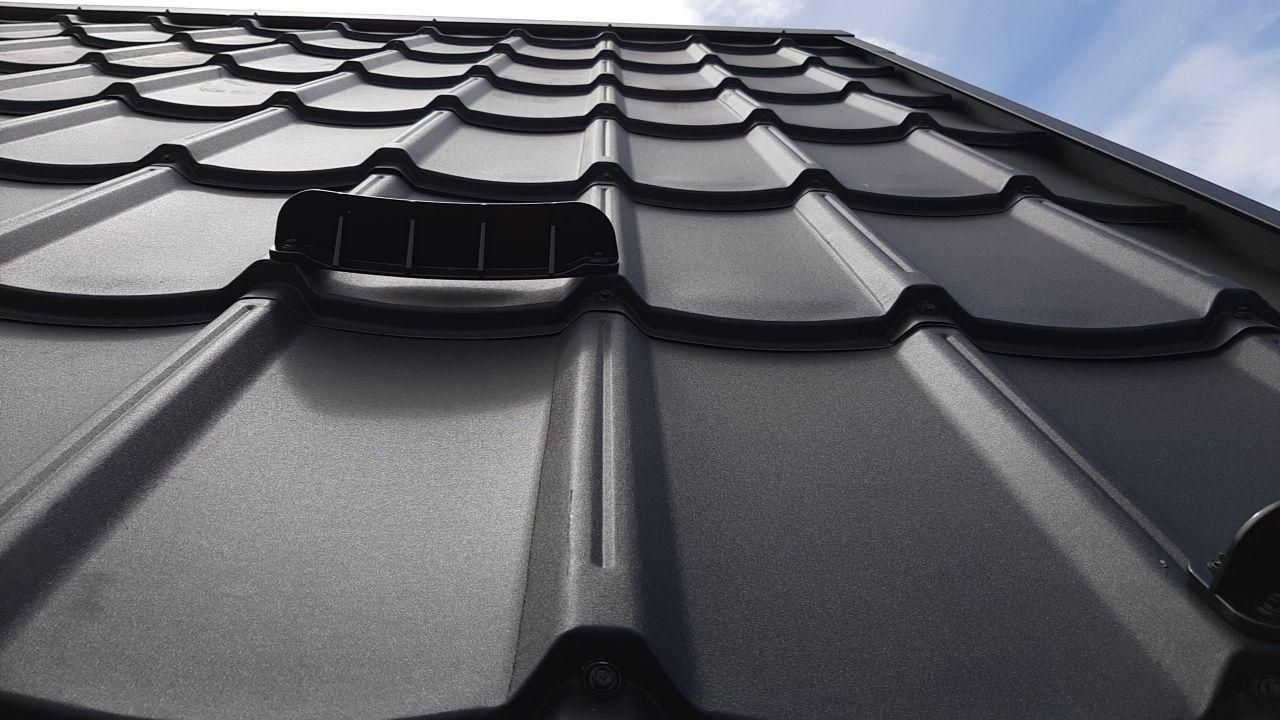 Close-up upward view of a dark gray metal tile roof with a vent attachment, showcasing the smooth reflective surface and interlocking design.