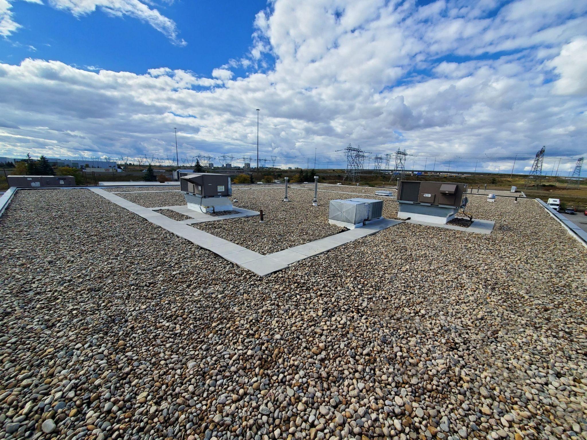Flat commercial roof covered with small round gravel ballast. HVAC units and other rooftop equipment are visible under a bright blue sky with scattered clouds.