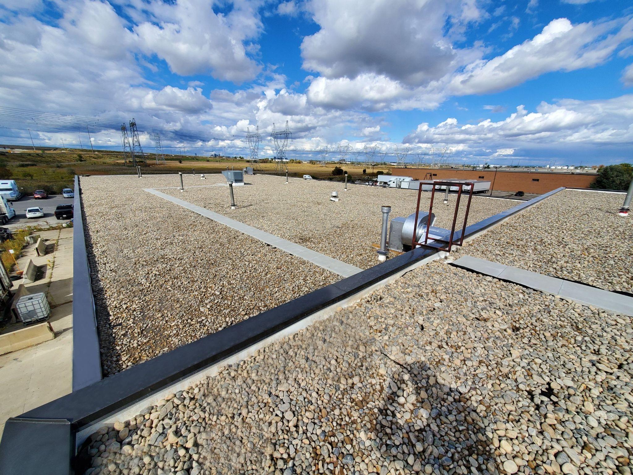 Flat gravel-ballasted roof of an industrial building with metal vents and rooftop equipment, viewed from above on a sunny day with blue sky and scattered clouds.