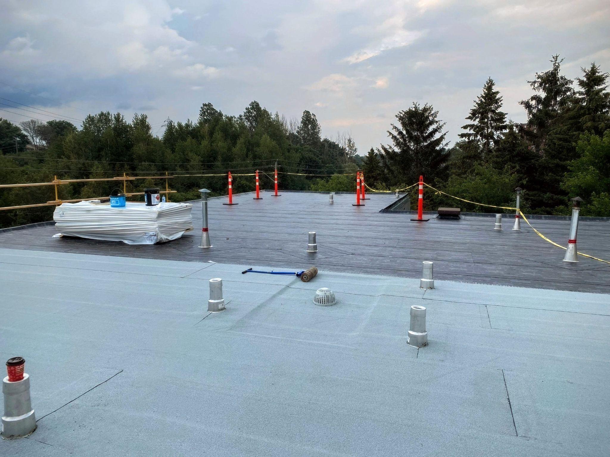 Flat roof installation in progress using modified bitumen material. Roofing rolls, ventilation stacks, and safety cones are arranged across the work area surrounded by trees.