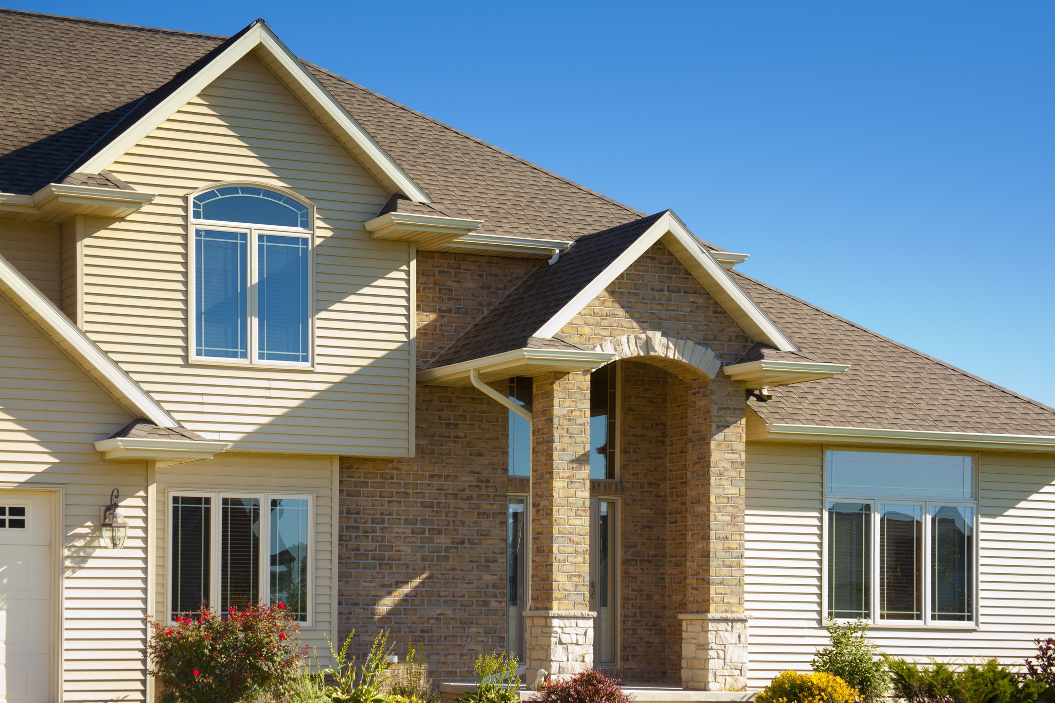 Suburban house with tan siding, gabled roof, and brick accents under a clear blue sky. Front garden features colorful shrubs and flowers.