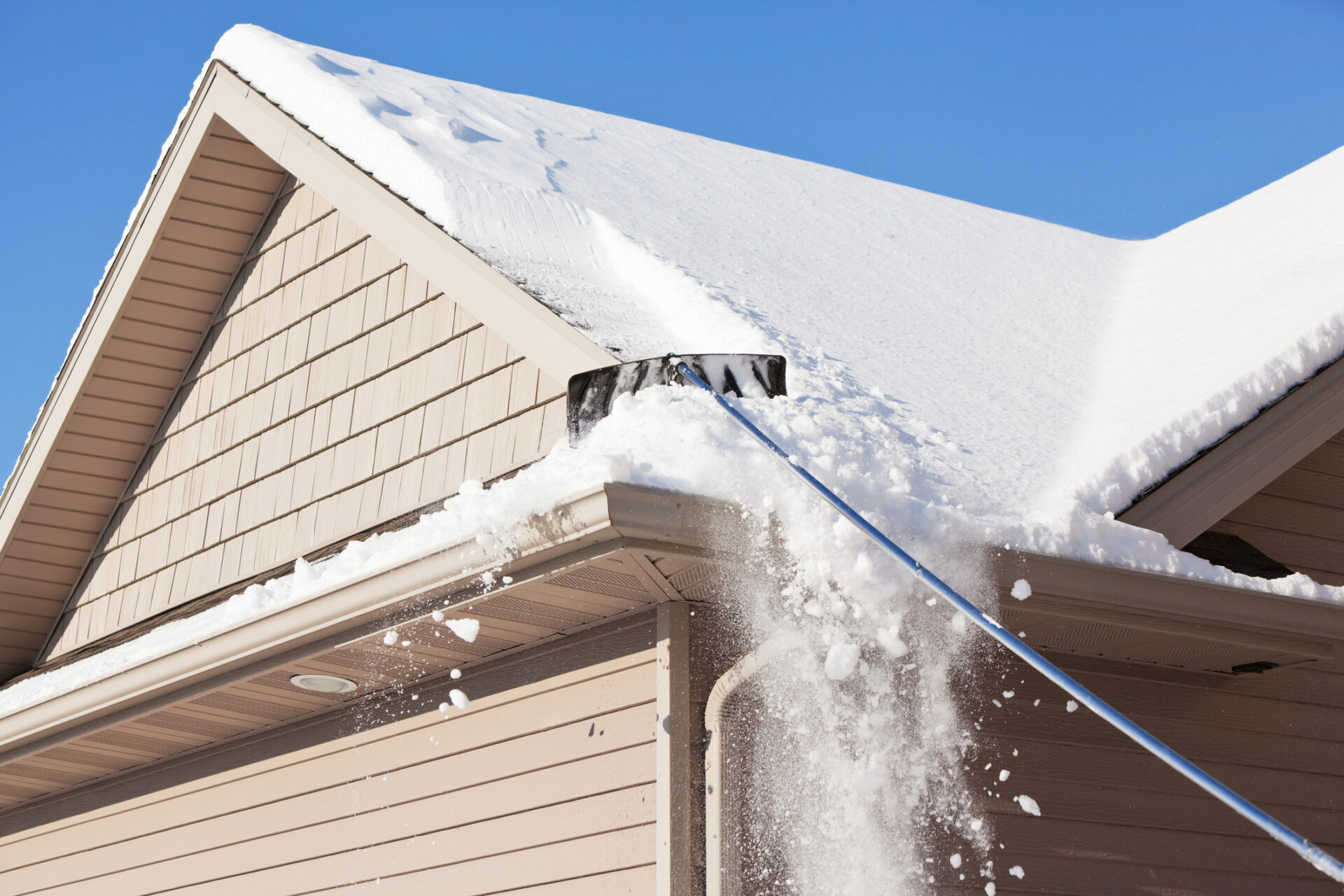 Person uses a roof rake to remove snow from a house, preventing buildup and potential damage. Bright blue sky in the background.