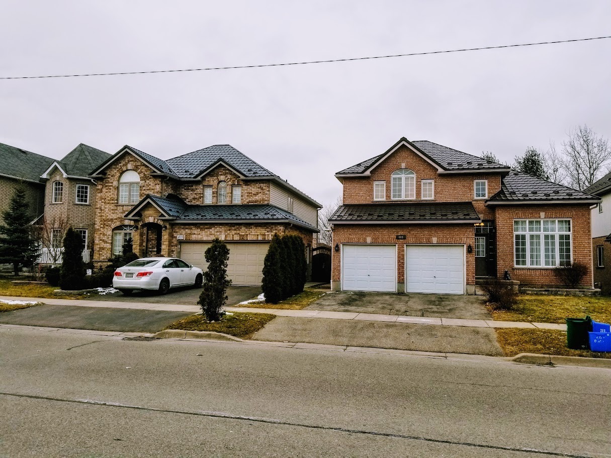 Suburban street with two brick houses featuring gabled roofs, each with a driveway and garage. A car is parked outside one house.