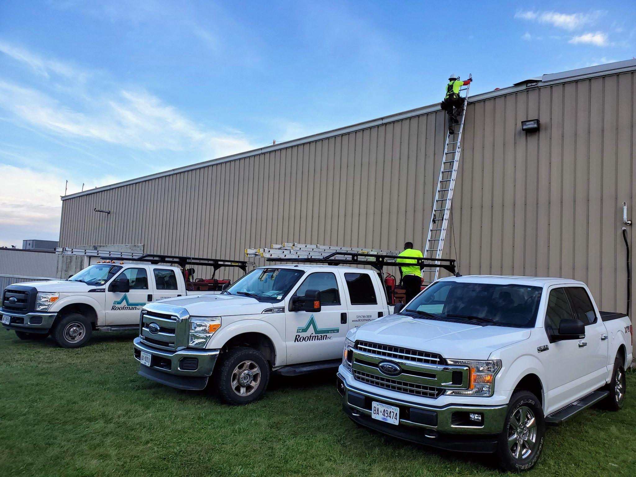 Three white work trucks labeled “Roofman” parked on a grassy area beside a beige industrial building. Workers are climbing ladders to access the roof under a clear blue sky.