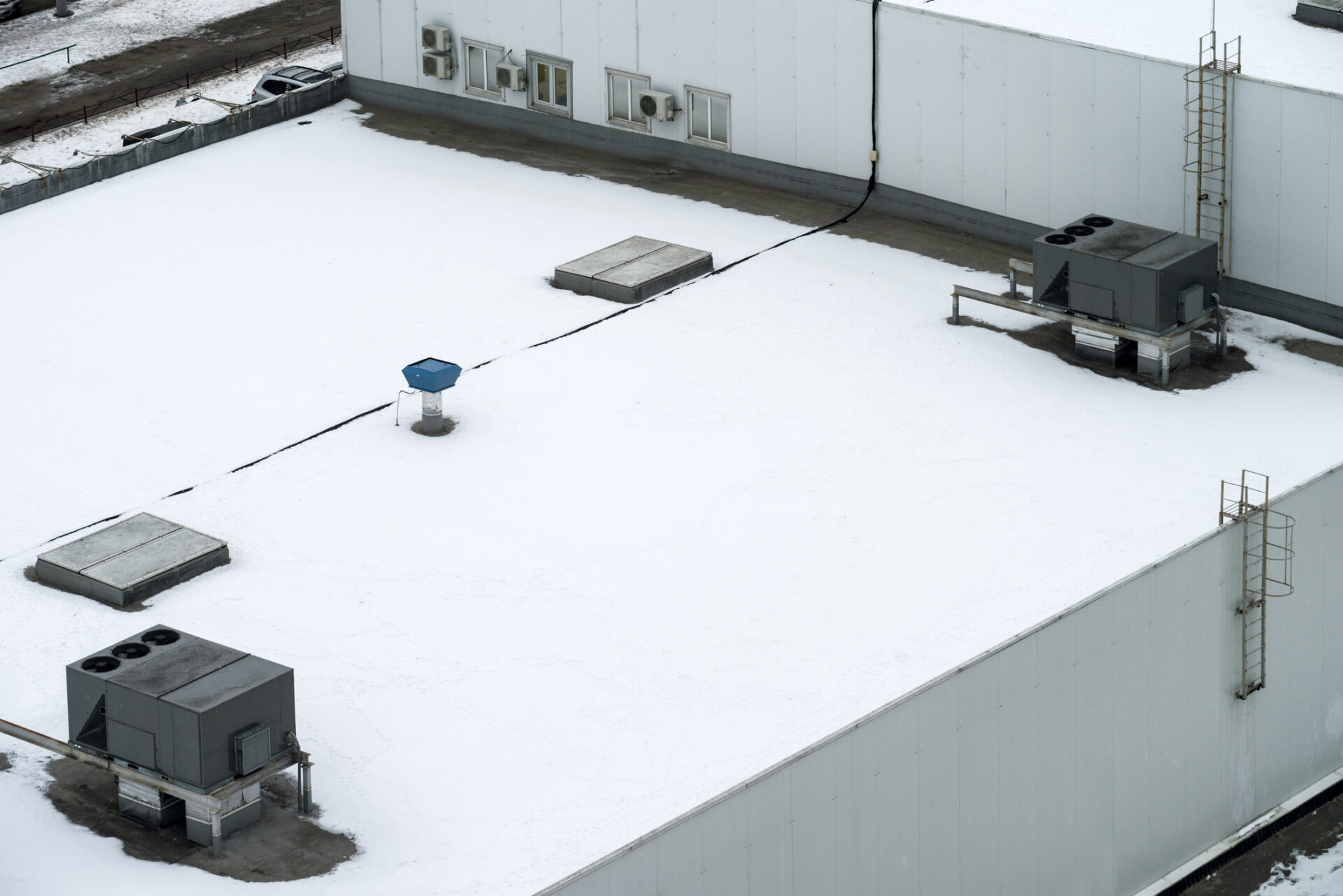 Snow-covered rooftop with air conditioning units and a ladder. No people or landmarks are visible; the setting appears industrial and minimalistic.