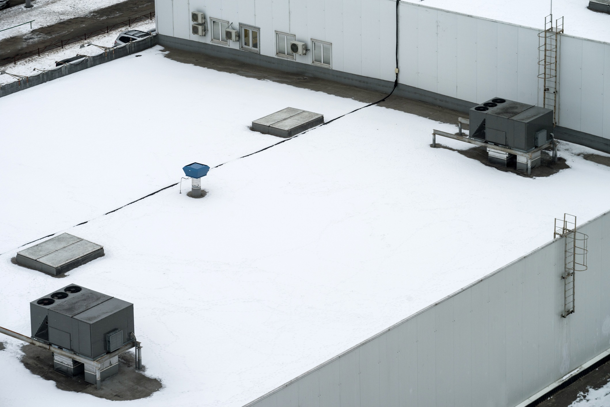 Snow-covered rooftop with air conditioning units and a ladder. No people or landmarks are visible; the setting appears industrial and minimalistic.
